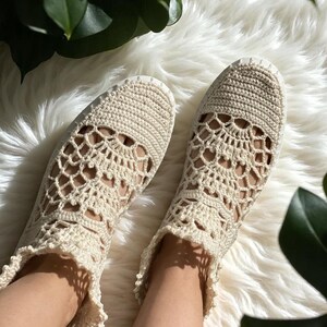 May include: A pair of cream-colored crocheted shoes with a delicate lace pattern. The shoes have a flat sole and are made with a soft, breathable material.
