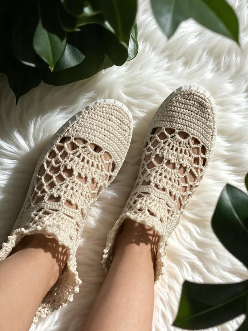 May include: A pair of cream-colored crocheted shoes with a delicate lace pattern. The shoes have a flat sole and are made with a soft, breathable material.