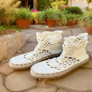 May include: A pair of white crocheted ankle boots with a delicate lace pattern. The boots have a flat sole and are made of a soft, lightweight material.