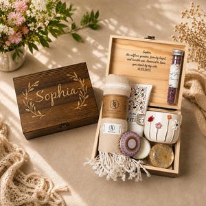 May include: A dark wood gift box engraved with the name Sophia and a leaf wreath, containing a beige fringed towel, a white floral mug, a glass tube of purple bath salts, a round patterned candle, and a circular stone, with a personalized message.