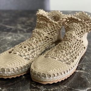 May include: A pair of beige crochet ankle boots with a white trim around the sole. The boots are made of a delicate, open-weave fabric.