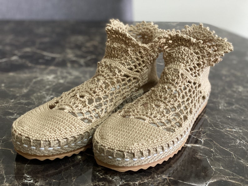 May include: A pair of beige crochet ankle boots with a white trim around the sole. The boots are made of a delicate, open-weave fabric.