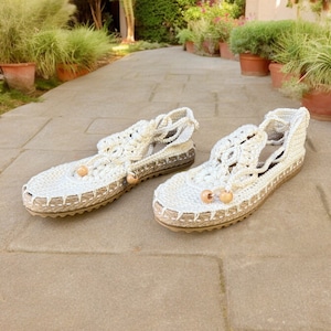 May include: A pair of white crocheted sandals with wooden beads. The sandals have a woven design and a flat sole.