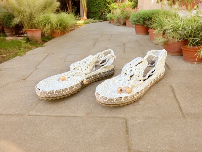 May include: A pair of white crocheted sandals with wooden beads. The sandals have a woven design and a flat sole.