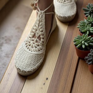 May include: A pair of white crocheted sandals with ankle ties and wooden beads. The sandals are on a wooden surface.