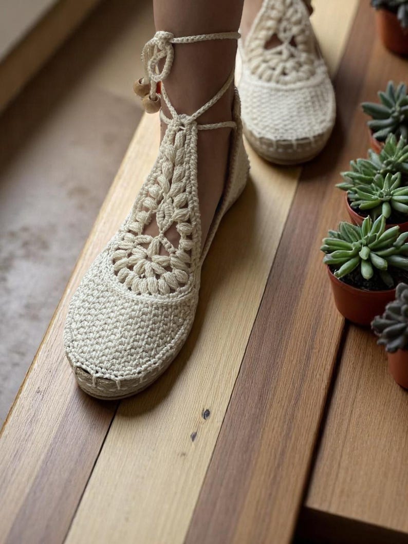 May include: A pair of white crocheted sandals with ankle ties and wooden beads. The sandals are on a wooden surface.