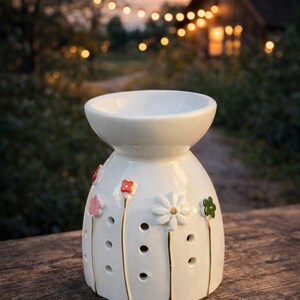 May include: White ceramic essential oil diffuser with floral accents. The diffuser has a round top and a bulbous base, decorated with red, pink, and green flowers. The base has several small holes. The background features a wooden surface and a blurred outdoor scene.