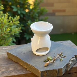 May include: A white ceramic essential oil diffuser with a rounded base and a bowl-shaped top. The diffuser has a small opening at the front and is placed on a dark gray stone. A small sprig of dried plant material and green herbs are next to the diffuser.