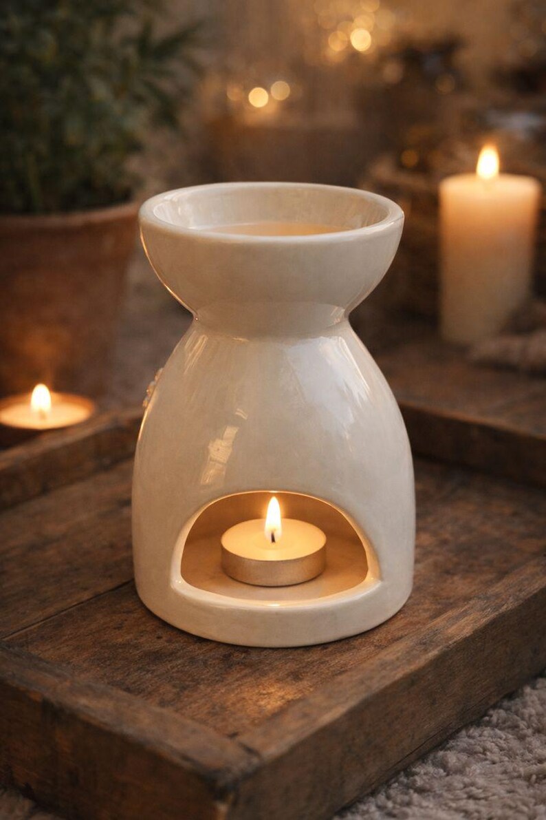 May include: Cream-colored ceramic essential oil diffuser with a lit tea light candle inside. The diffuser sits on a wooden tray, with a lit candle in the background. The design is simple and elegant, perfect for aromatherapy.