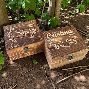 May include: Two wooden boxes with hinged lids and metal clasps. The boxes are stained a dark brown and feature the names "Steph" and "Cristina" in a decorative font, surrounded by floral designs. The boxes are sitting on the ground.