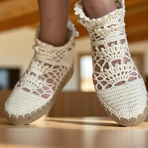 May include: A pair of white crocheted espadrille shoes with a delicate lace pattern. The shoes have a ruffled trim around the ankle and a jute sole.