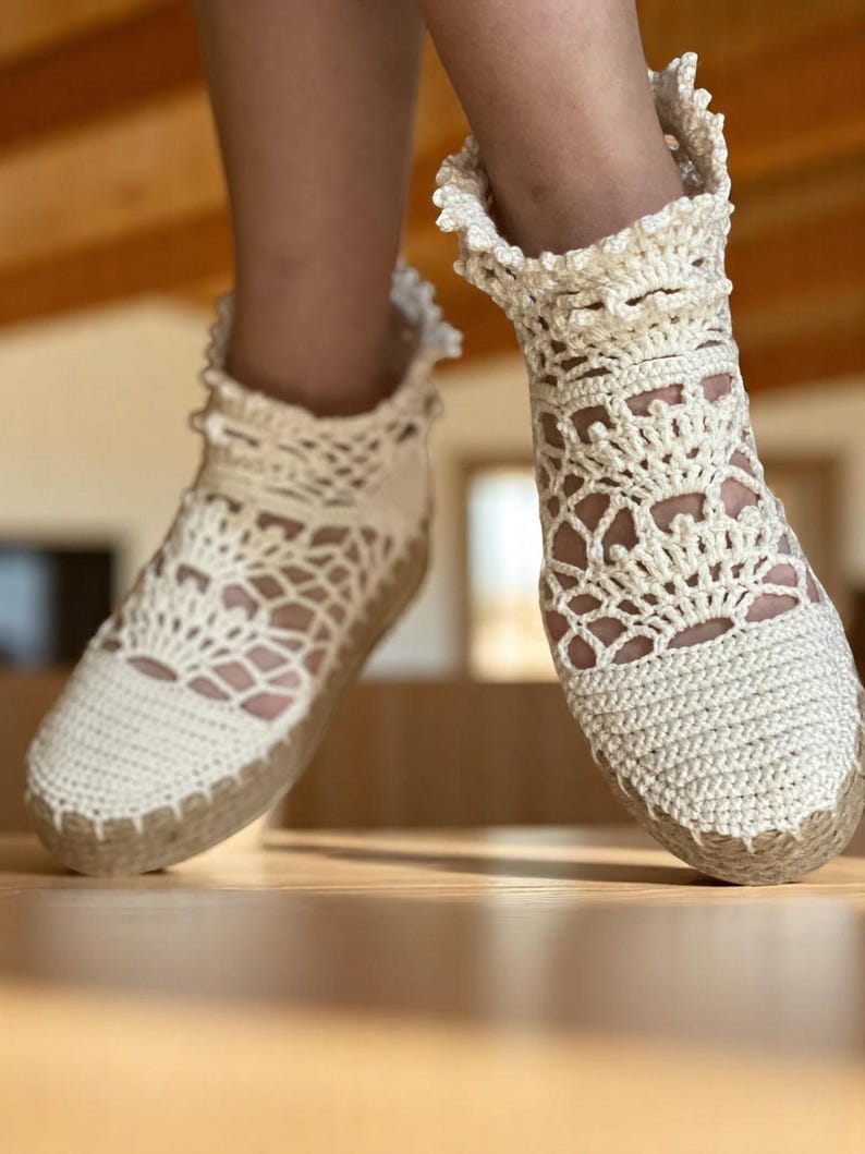 May include: A pair of white crocheted espadrille shoes with a delicate lace pattern. The shoes have a ruffled trim around the ankle and a jute sole.