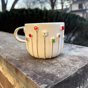 May include: White ceramic mug with a gold handle and rim, adorned with colorful, raised flower designs. The flowers are red, pink, and green, with gold stems. The mug is sitting on a stone surface.