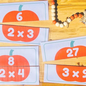 May include: Educational puzzle cards featuring orange pumpkin shapes with numbers and multiplication problems. The cards are designed to teach basic math skills. A wooden bead garland and a tag that says "Give thanks" are also visible.