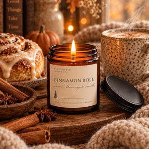 May include: A lit candle in an amber glass jar with a cream-coloured label reading NICOLAYSEN CANDLE CO, CINNAMON ROLL, cinnamon brown sugar vanilla, and 4 OZ | HAND POURED IN OREGON. The jar has a black lid resting beside it on a wooden surface.