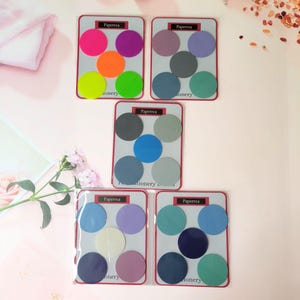 Peut inclure: Cinq ensembles d'autocollants ronds colorés avec le texte "Paperrca Stationery" sur chaque carte. Les autocollants sont dans différentes nuances de bleu, vert, violet, rose et orange.