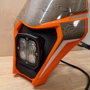 May include: An orange motorcycle headlight assembly with a patterned visor. The headlight features a black housing with four LED lights and the brand name "DENALI". The visor has a brown and gray wood grain pattern.