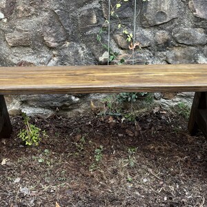 May include: A long wooden bench with a dark brown stain. The bench has a simple design with two A-frame legs.
