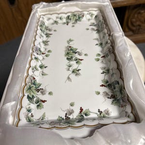 May include: A white porcelain serving tray with a scalloped edge and a floral design featuring green leaves and red berries. The tray is in a white box with a satin lining.