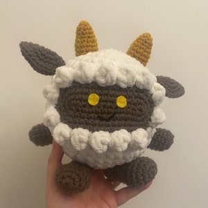 May include: A crocheted plush toy shaped like a sheep with brown legs and horns. The sheep has a brown face with yellow eyes and a white fluffy body.