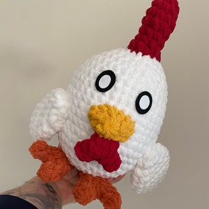 May include: A white crocheted chicken plush toy with red, orange, and yellow accents. The chicken has a red comb, orange feet, and a yellow beak.