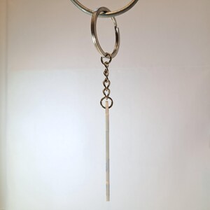 Puede incluir: Un llavero plateado con una peque&ntilde;a etiqueta rectangular blanquecina. La etiqueta est&aacute; suspendida de una cadena y un llavero partido. El llavero cuelga sobre un fondo blanco.