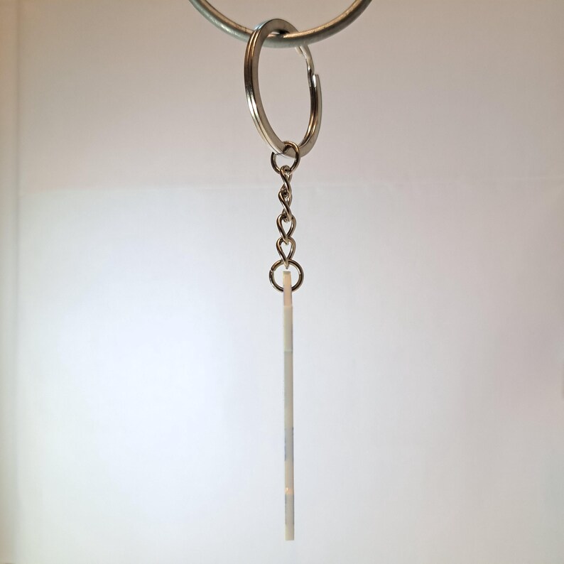 Puede incluir: Un llavero plateado con una peque&ntilde;a etiqueta rectangular blanquecina. La etiqueta est&aacute; suspendida de una cadena y un llavero partido. El llavero cuelga sobre un fondo blanco.