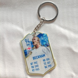 May include: A football-themed keychain with a card design. The card features a photo of Beckham, with stats including 87 PAC, 90 SHO, and 97 PAS. The keychain has a silver ring and chain.