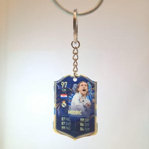 Puede incluir: Un llavero azul y dorado con la imagen y las estad&iacute;sticas de un futbolista. El llavero tiene una cadena plateada y un colgante en forma de escudo. El jugador se llama Modrić, y la tarjeta muestra sus estad&iacute;sticas.