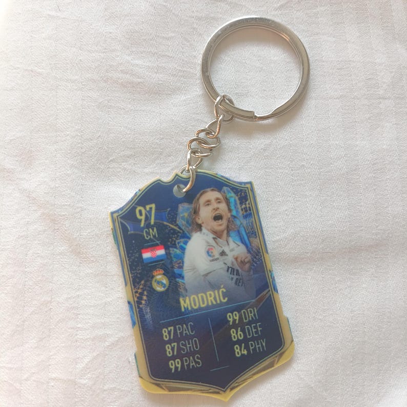 Puede incluir: Un llavero con un dise&ntilde;o azul y dorado, con la imagen y las estad&iacute;sticas de un jugador de f&uacute;tbol. El llavero tiene un anillo y una cadena de metal plateado. El jugador es Modrić, y la tarjeta muestra 97 cm.