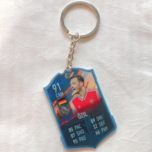 May include: A blue and white keychain featuring a soccer player's image and stats. The keychain has a silver ring and chain. The player's name is Özil, with stats including 85 PAC, 87 SHO, and 95 PAS.