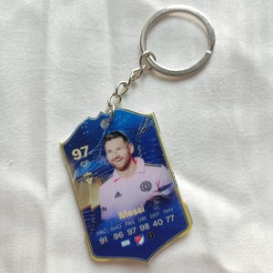 Op de afbeelding: Een blauw en gouden sleutelhanger met een foto van een voetballer. De sleutelhanger heeft een schildvorm met het nummer 97 en de naam Messi. De sleutelhanger is bevestigd aan een zilveren metalen ring.