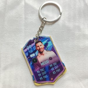 Op de afbeelding: Sleutelhanger met een zilverkleurige ring en ketting, met de afbeelding van een voetballer. De kaartvormige hanger toont een foto van een voetballer, de naam "MESSI" en spelersstatistieken. De achtergrond is een verloop van paars en blauw.
