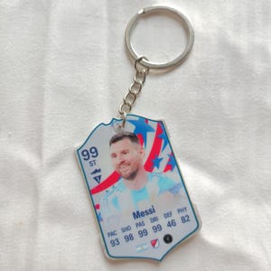 May include: A keychain featuring a soccer player's image on a card-shaped pendant. The card has a blue and white design with the player's photo, name, and statistics. The keychain includes a silver-colored ring and chain.