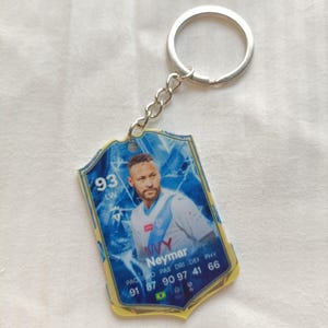 May include: A keychain featuring a soccer player, Neymar, with a blue and yellow design. The keychain has a silver key ring and chain. The card-shaped pendant displays player stats and the number 93.