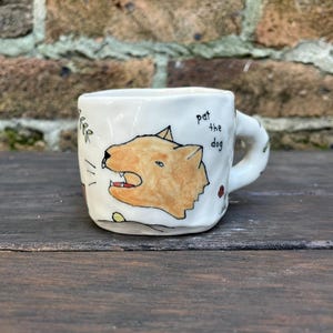 Może przedstawiać: Mały, kremowy ceramiczny kubek z ręcznie malowaną ilustracją warczącego, pomarańczowo-brązowego psa. Na kubku napis "pat the dog" w kolorze czarnym. Uchwyt jest zaokrąglony, z małymi kwiatowymi akcentami.