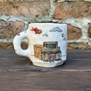 Könnte beinhalten: Eine weiße Keramiktasse mit handgemaltem Design eines Gebäudes mit einer roten Tür, Wolken und einem roten Vogel, der am Himmel fliegt.