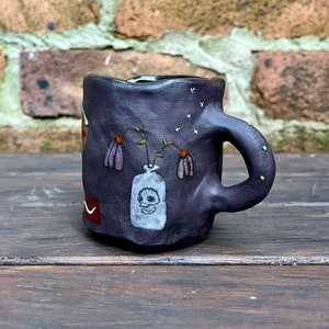 May include: A dark purple ceramic mug with a textured surface. The mug features a hand-painted design of a vase with a skull and flowers, along with stars. The handle has small yellow accents. The mug is sitting on a wooden surface.
