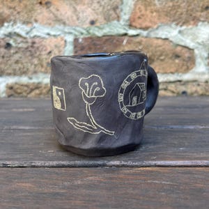 May include: A small, dark brown ceramic mug with a handle. The mug has a white design on it, including a flower, a house, and a geometric shape.
