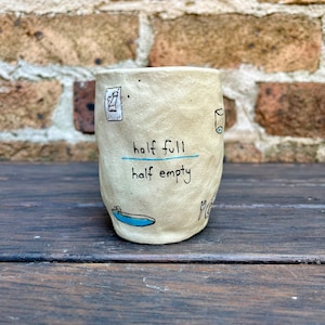 Puede incluir: Una taza de cerámica blanca con ilustraciones dibujadas a mano en negro y el texto "half full half empty".