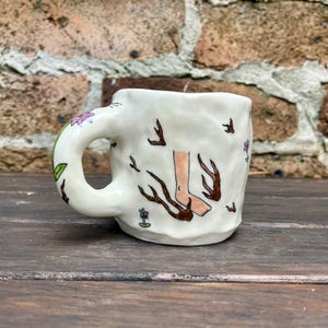 May include: White ceramic mug with a hand-painted design of a leg and foot walking on branches. The mug has a brown handle and is decorated with small pink flowers.