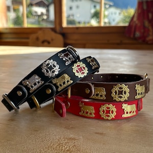 Op de afbeelding: Drie leren hondenhalsbanden met decoratieve metalen koe- en bloemenmotieven. De halsbanden zijn zwart, bruin en rood.