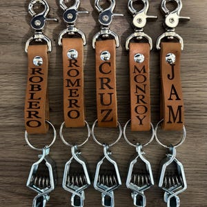 May include: Five personalized leather keychains with silver-tone hardware. Each keychain features a brown leather strap engraved with a name: ROBLEO, ROMERO, CRUZ, MONROY, and JAM. The keychains have a spring clip and a split ring.