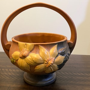 May include: A brown ceramic bowl with a handle, decorated with yellow and brown flowers. The bowl has a dark brown base.