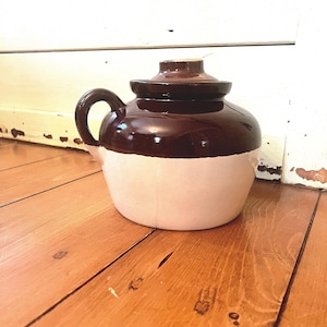 Peut inclure: Une cruche en céramique brune et blanche avec un couvercle et une anse. La cruche est émaillée et a un aspect rustique.