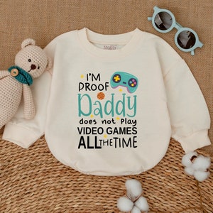 May include: A cream colored baby onesie with a green and blue video game controller graphic and the text "I'm Proof Daddy does not play video games all the time".