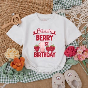 May include: White short-sleeved onesie with red and white checkered fabric and red strawberries. The onesie has the text "Olivia's Berry 1st Birthday" printed on it.