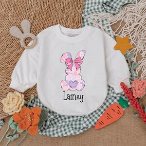 May include: White long-sleeved shirt with a pink and purple bunny with a pink bow and a heart on its chest. The shirt has the name "Lainey" printed below the bunny.