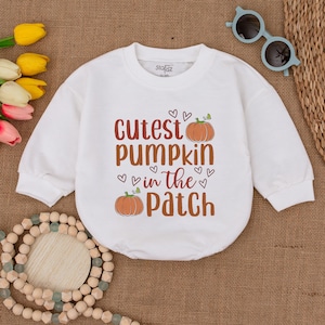 May include: White long-sleeve onesie with the text "Cutest pumpkin in the patch" and a small orange pumpkin graphic. The onesie has a crew neck and is decorated with small hearts.