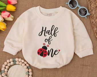 May include: A cream coloured long-sleeved onesie with the text "Half of one" and a red ladybug with black spots and red flowers.